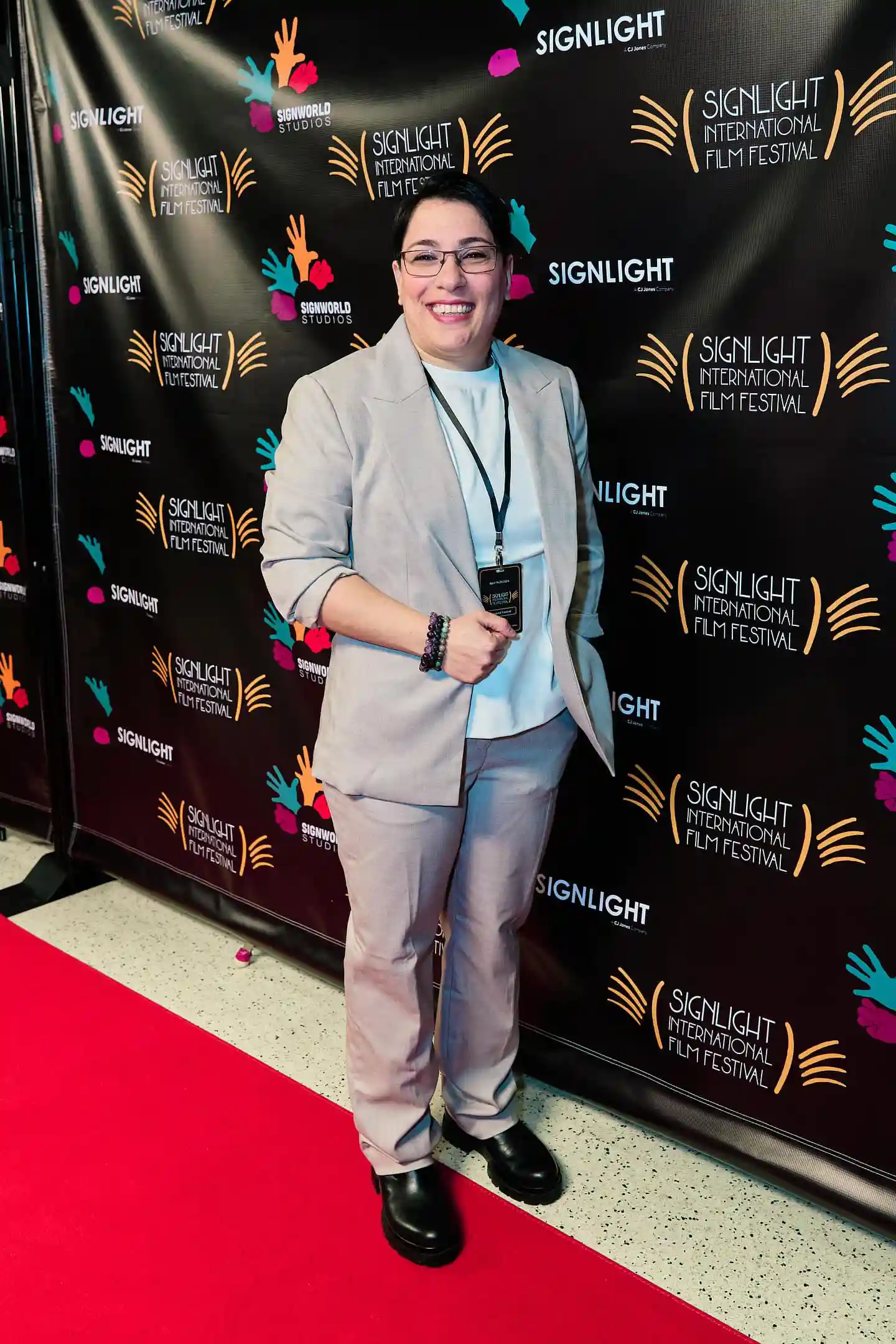 A person with short dark hair and glasses stands on a red carpet at the SignLight International Film Festival. They are dressed in a light beige suit, a white top, and black shoes, smiling broadly. The backdrop behind them has the festival's colorful logo and branding.