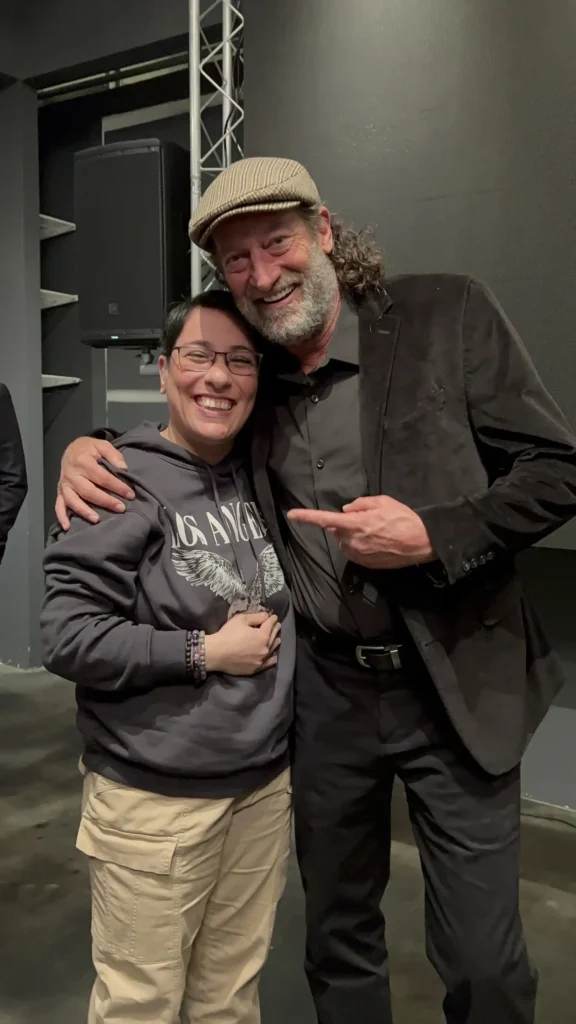 A person with short dark hair and glasses, wearing a dark hoodie with "Los Angeles" printed on it and beige cargo pants, smiles widely while standing next to a man with curly gray hair, a beard, and a flat cap. The man is pointing toward them, and both appear to be in a casual indoor setting, possibly at an event. The two people are Ladan Sahraei and Troy Kotsur, Oscar winner for Best Supporting Actor for CODA copy