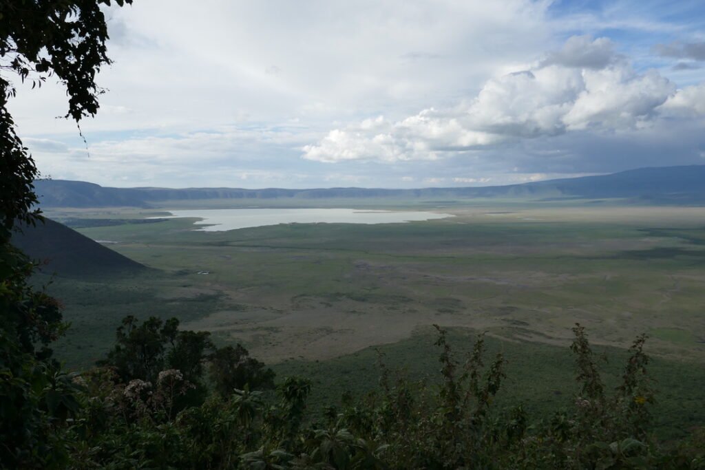 Scenic view of Ngorongoro Crater, showcasing expansive green plains, a reflective lake in the distance, and surrounding highlands under a partly cloudy sky