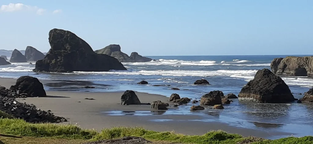 A scenic coastal landscape featuring large, rugged rock formations scattered along the sandy shoreline. Waves gently crash against the rocks, with the deep blue ocean stretching into the horizon under a clear sky. Green vegetation borders the foreground, adding contrast to the dark rocks and shimmering water - Rocky shores of the Oregon Trail copy