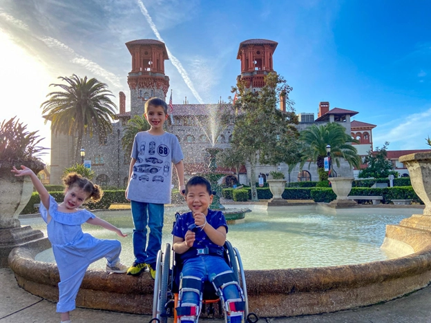 Three smiling children pose in front of a beautiful fountain with an ornate historic building and palm trees in the background. The child on the left, wearing a white outfit, strikes a playful pose with an arm raised. The child in the middle, wearing a gray T-shirt and jeans, stands confidently. The child on the right, seated in a wheelchair, wears a blue outfit and beams with joy. The scene is bathed in warm sunlight, with a clear blue sky overhead.