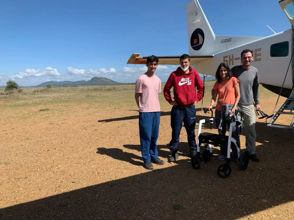 A group of five people standing next to a small airplane on a dirt airstrip under a clear blue sky. One of the individuals is using a walker for mobility. In the background, there are rolling hills and open grasslands.