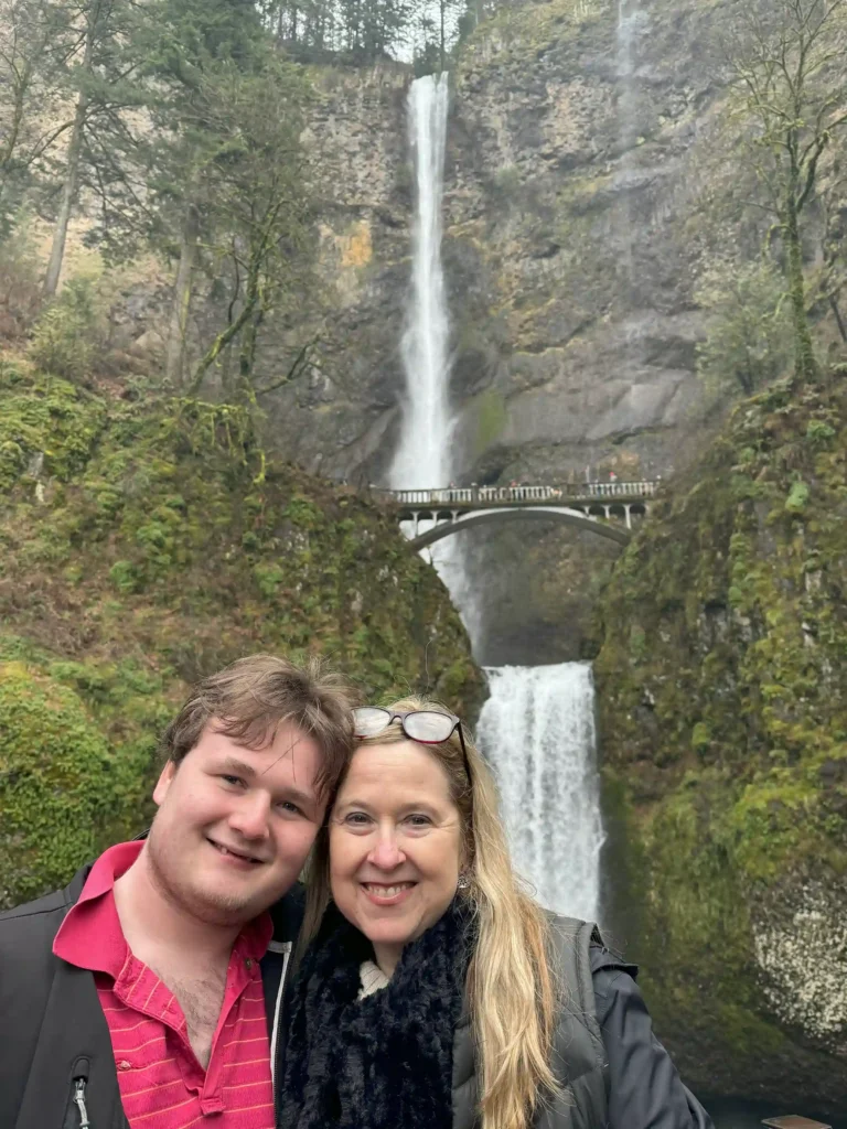 Two people smiling in front of Multnomah Falls, a stunning two-tiered waterfall surrounded by lush greenery and moss-covered cliffs. A historic bridge spans across the lower section of the waterfall, creating a picturesque scene."