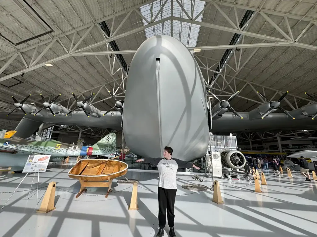 Person standing in front of the massive nose of the Spruce Goose airplane inside a museum hangar, showcasing its enormous size and the scale of its 319-foot wingspan. Other aircraft and exhibits are visible in the background, emphasizing the impressive engineering of the Spruce Goose.