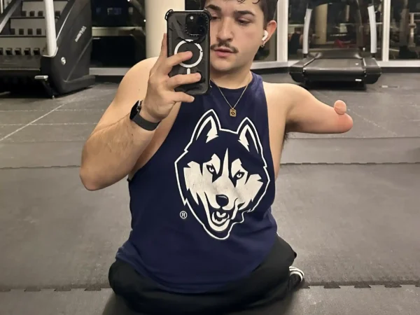 A man in a gym takes a selfie in front of a mirror. He has short, curly hair, a mustache, and is wearing a navy blue tank top with a husky logo. One of his arms ends above the elbow. In the background, there are treadmills and large windows, showcasing a nighttime urban view.
