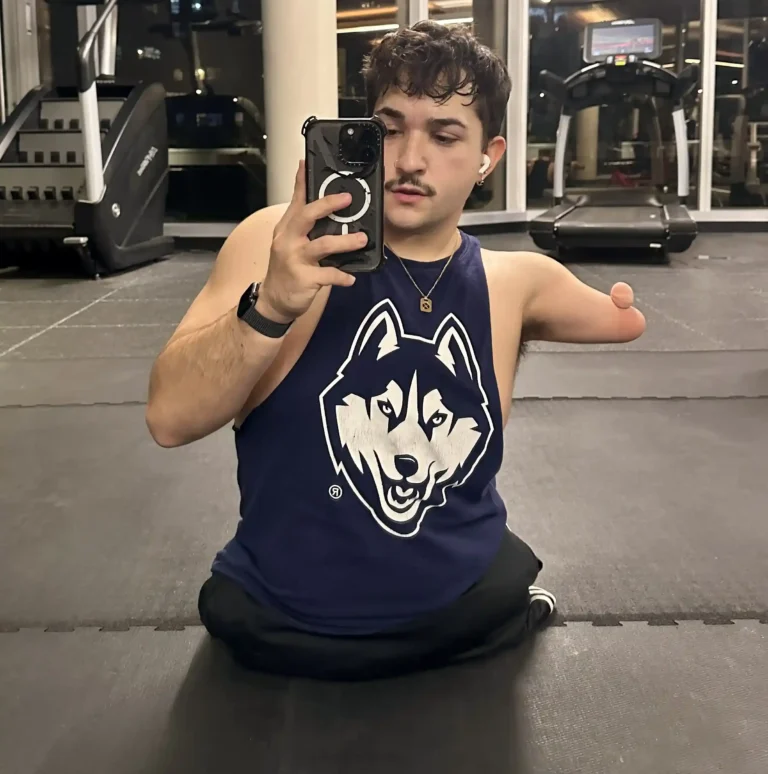 A man in a gym takes a selfie in front of a mirror. He has short, curly hair, a mustache, and is wearing a navy blue tank top with a husky logo. One of his arms ends above the elbow. In the background, there are treadmills and large windows, showcasing a nighttime urban view.