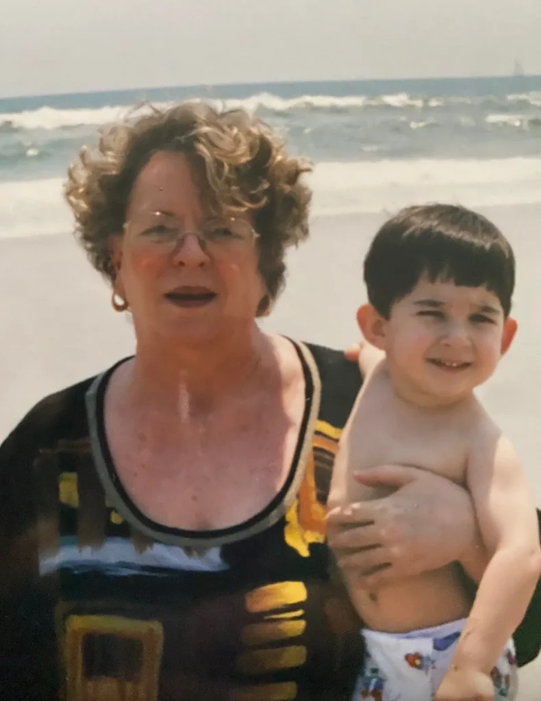 An older woman with curly hair, wearing glasses and a patterned top, is holding a young child at the beach. The child has dark hair and is wearing a diaper, with the ocean waves visible in the background.