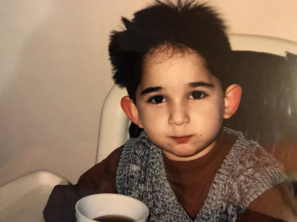 This is a photograph of a young child with dark hair wearing a brown shirt and a knitted vest. The child is sitting in a high chair, holding a cup. The background is simple and out of focus, emphasizing the child’s facial expression and surroundings.