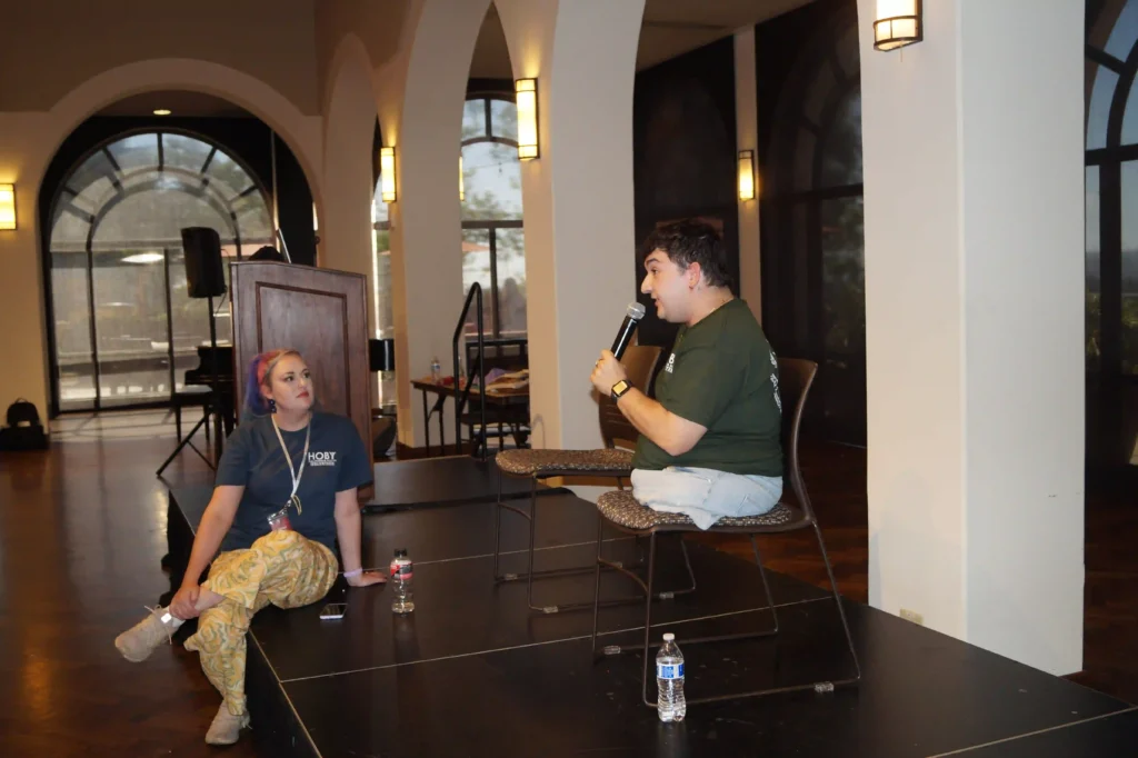Two individuals engage in a conversation at an indoor event. The person on the left, seated on the edge of a platform, listens attentively, wearing a blue shirt with a logo and patterned yellow pants. The individual on the right, seated on a chair on the stage, speaks into a microphone, wearing a green shirt and jeans. He has one arm only and no legs. The setting features large arched windows, soft lighting, and a podium in the background