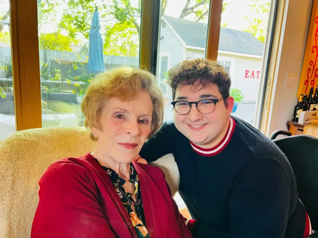 An elderly woman with short, light-colored hair wearing a maroon cardigan and floral blouse sitting on a beige chair, smiling warmly next to a young man with dark curly hair, glasses, and a navy sweater with red and white stripes, both posing together indoors near a large window with natural light streaming in. The background shows a backyard with greenery, a small shed, and outdoor furniture.