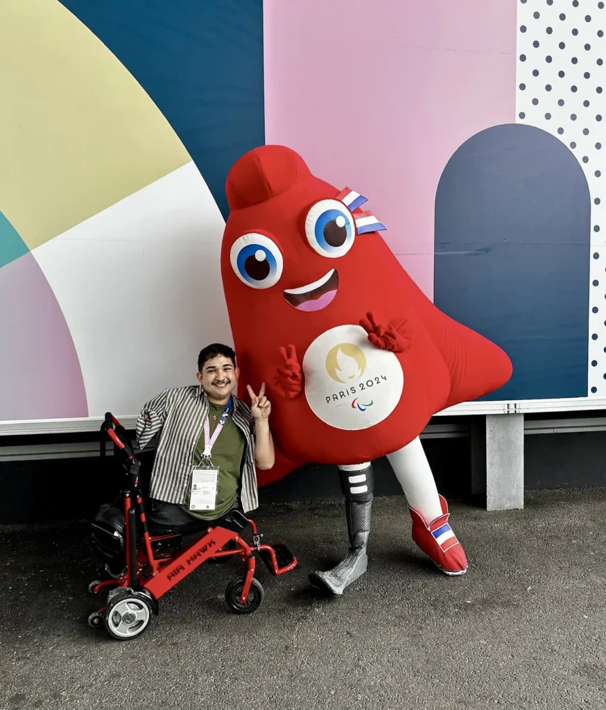 A person seated in a red mobility aid, smiling and making a peace sign, poses next to the official Paris 2024 Paralympic mascot. The mascot is red, shaped like a Phrygian cap with a prosthetic leg and large, friendly eyes, standing in front of a colorful geometric background.