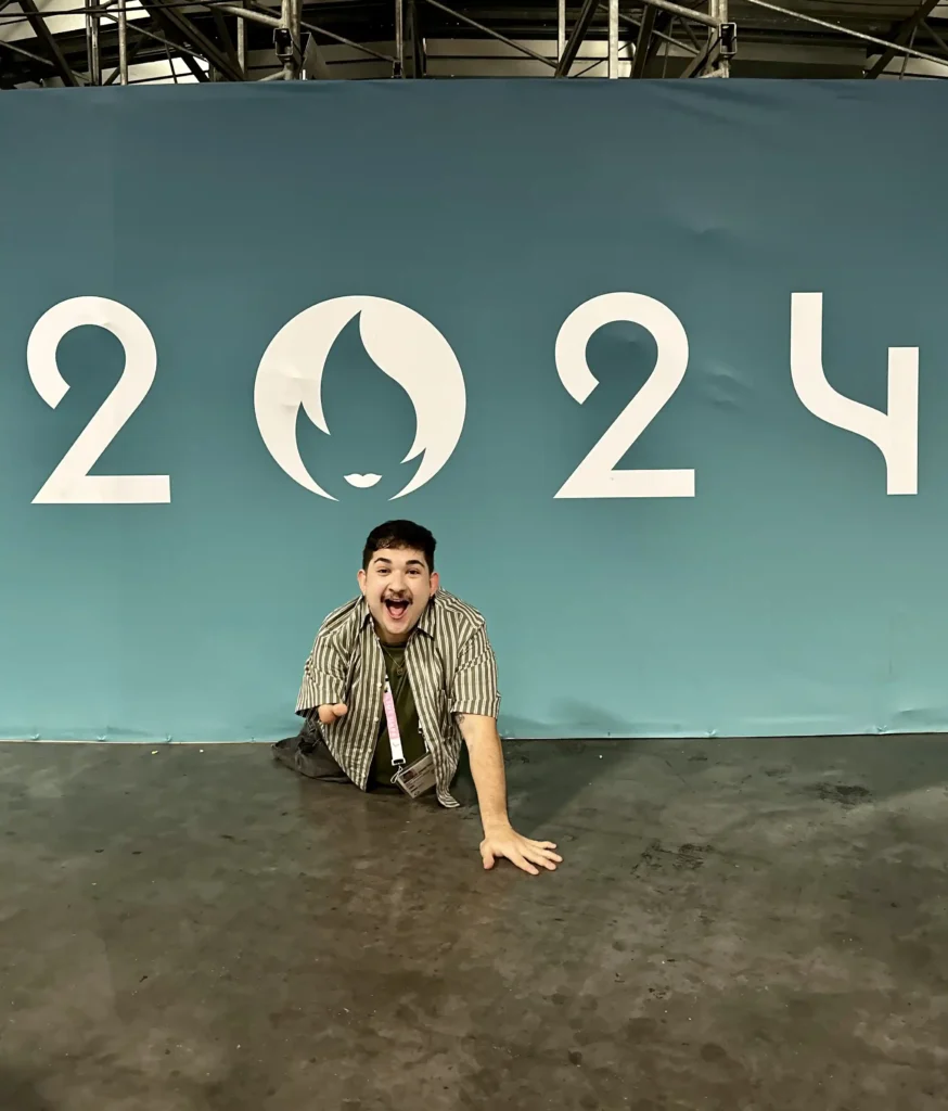 A man with a joyful expression and short dark hair is positioned in front of a large blue backdrop with the number "2024" prominently displayed. The backdrop features a stylized flame or abstract logo between the "2" and "4." The man, wearing a striped shirt and lanyard, leans forward enthusiastically, resting on one arm and the floor. The setting appears to be an event or convention space.
