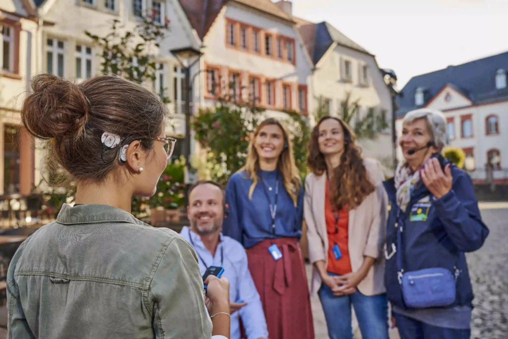 A group of people standing on a cobblestone street in a European town, with colorful buildings in the background. A woman in the foreground with a cochlear implant and glasses is facing the group, holding a small device. The group, including an older woman with a headset and three younger adults, is smiling and engaging with the woman