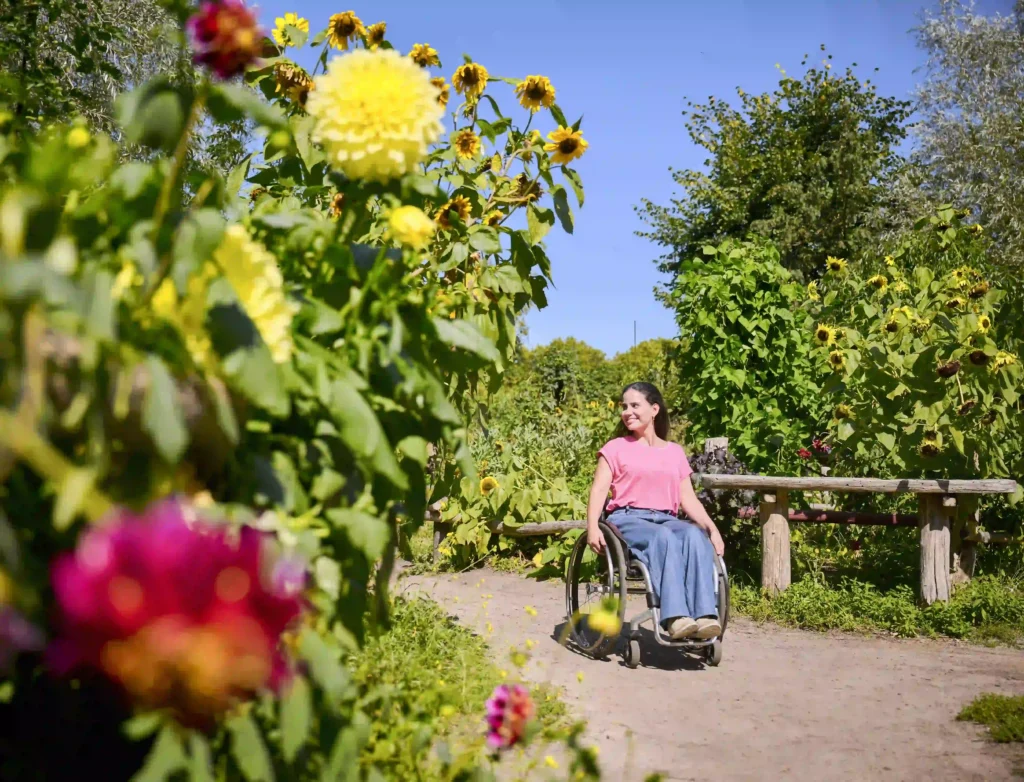 A person in a wheelchair enjoying a sunny day in a vibrant garden, surrounded by tall sunflowers and colorful flowers, with a clear blue sky in the background. The individual is smiling and appears relaxed on a dirt pathway next to a rustic wooden bench.