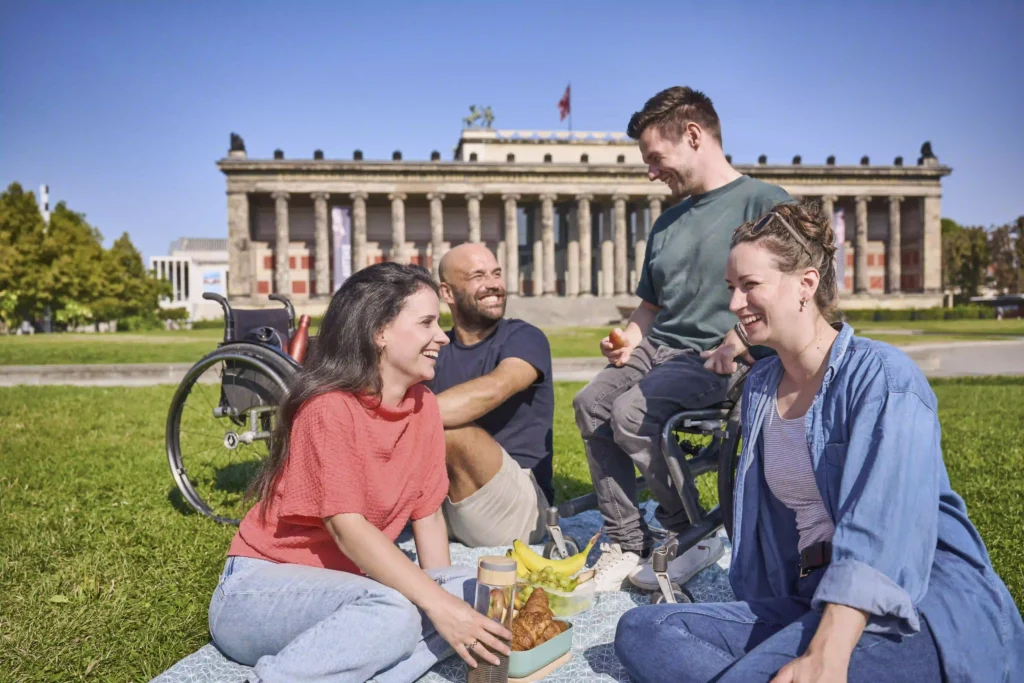 A group of four friends, including a person in a wheelchair, enjoying a picnic on a sunny day in front of a historic building with columns and statues on top. They are seated on a blanket with food items like croissants, bananas, and grapes. The atmosphere is cheerful, with everyone smiling and engaging in conversation. Lush green grass and a clear blue sky enhance the vibrant and inclusive setting.