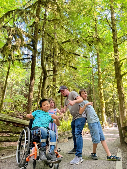 A group photo taken on a forest trail surrounded by tall trees covered in moss. The group includes a child in a wheelchair wearing a blue shirt, a man wearing a hat and sunglasses making playful poses, and two other children smiling and striking fun poses. The scene is lit with natural sunlight filtering through the dense green canopy above, creating a serene and joyful atmosphere.