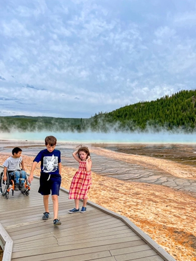 Three children walking on a boardwalk in front of a steaming geothermal hot spring in Yellowstone National Park. The child on the left is in a wheelchair, the boy in the middle is wearing a blue t-shirt and shorts, and the girl on the right is wearing a red checkered dress and holding her hair against the wind. The background features mist rising from the colorful hot spring and a lush green forest.