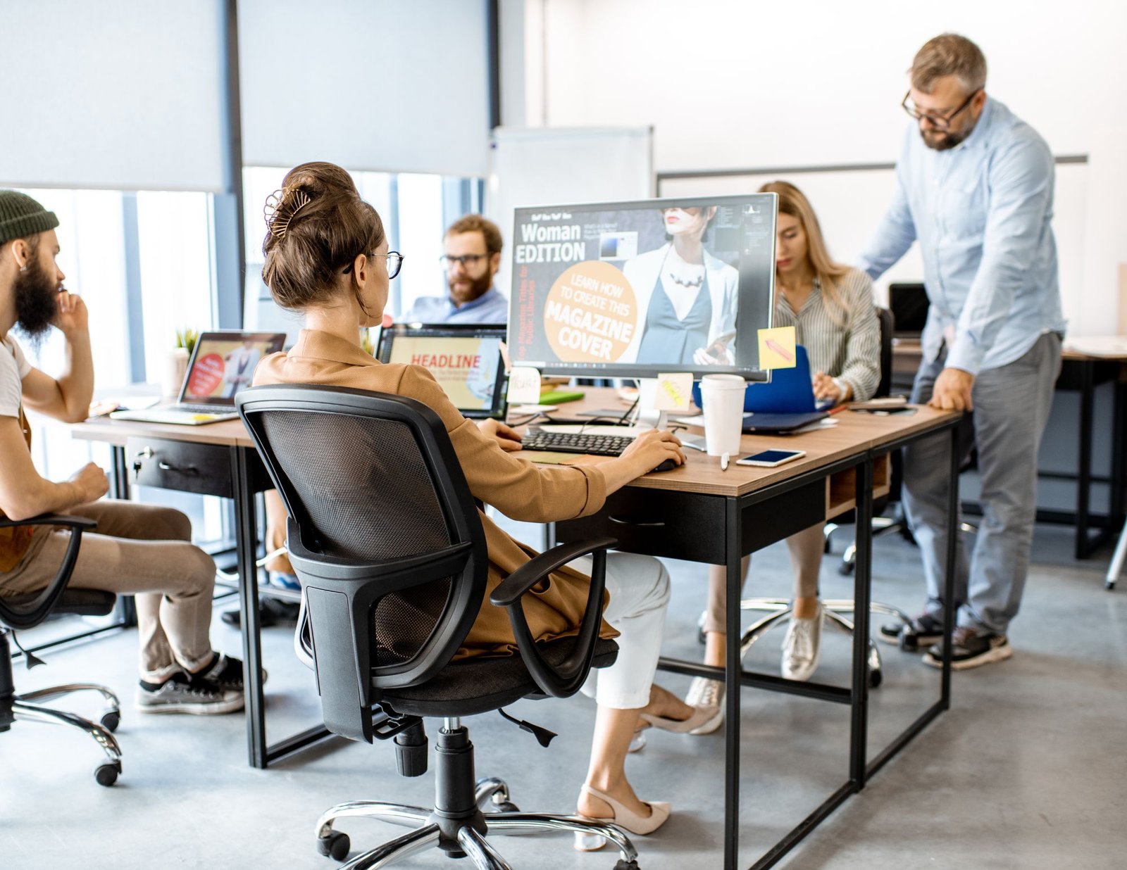 A team of professionals collaborating in a modern office space, focused on designing a magazine cover displayed on a computer screen. A woman sits at the forefront, working on the design, while other team members engage in discussion around the table. Laptops, notes, and coffee cups are scattered on the desks, creating an energetic and creative workspace atmosphere.