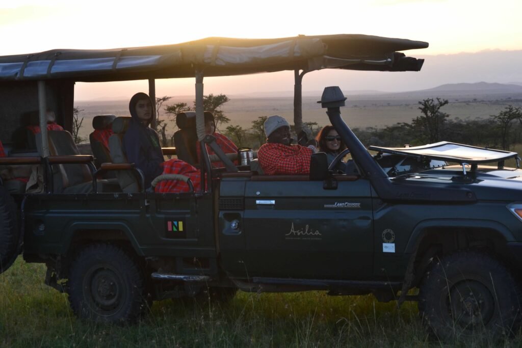 Safari vehicle at sunset with passengers enjoying a game drive, parked in the middle of a grassy savanna landscape. The vehicle, a dark green Land Cruiser branded with 'Asilia,' features red Maasai blankets draped over the seats. A guide in traditional Maasai attire is seated at the front, along with other passengers wearing jackets, smiling and observing the surroundings. The horizon shows acacia trees and rolling plains under a glowing golden sunset.