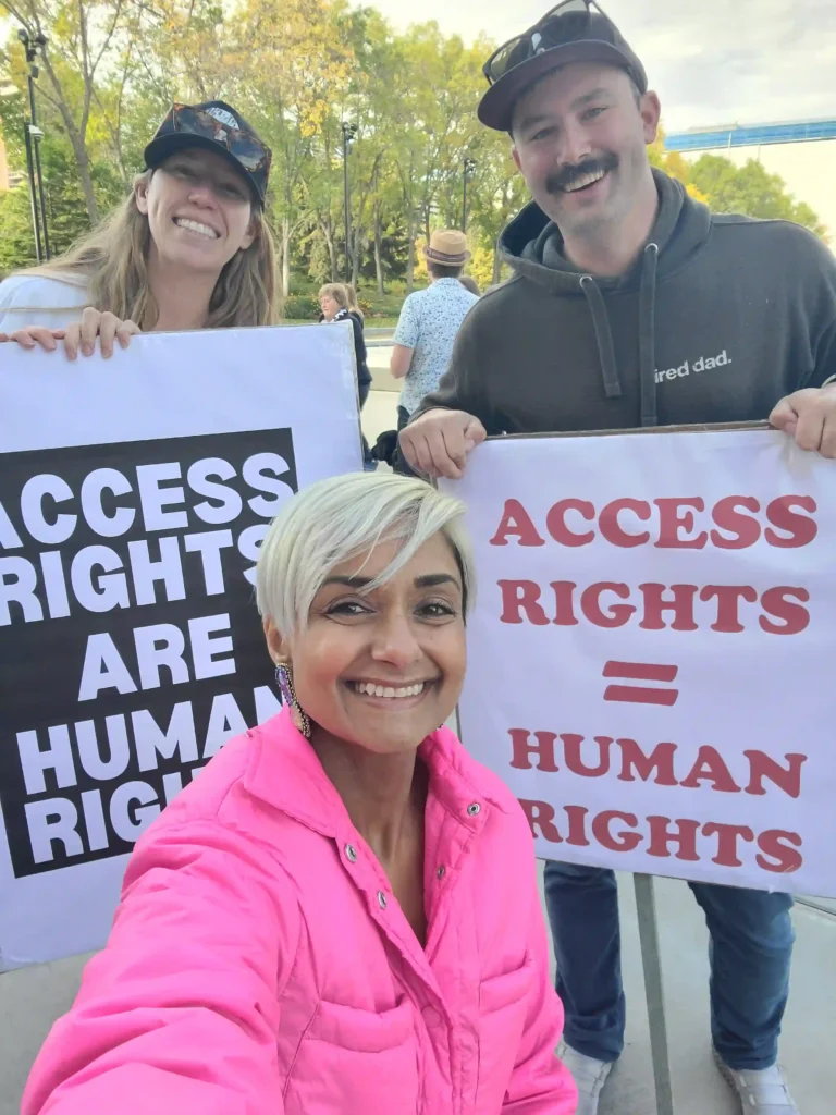 A group of three people smiling and holding signs advocating for accessibility rights. The signs read, "ACCESS RIGHTS ARE HUMAN RIGHTS" and "ACCESS RIGHTS = HUMAN RIGHTS." The person in the foreground, taking a selfie, is wearing a bright pink jacket and has short, platinum blonde hair.