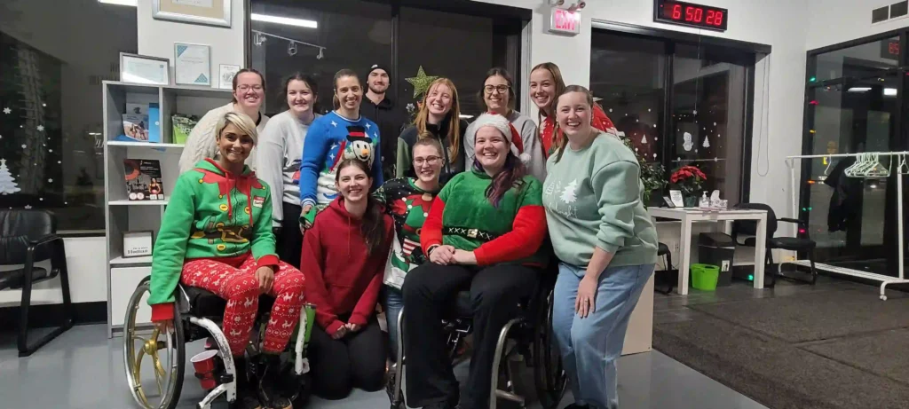 : A group of people gathered in a festive indoor setting, dressed in Christmas-themed sweaters and attire. Two individuals in the group are using wheelchairs. The environment includes holiday decorations, bookshelves, and a welcoming atmosphere