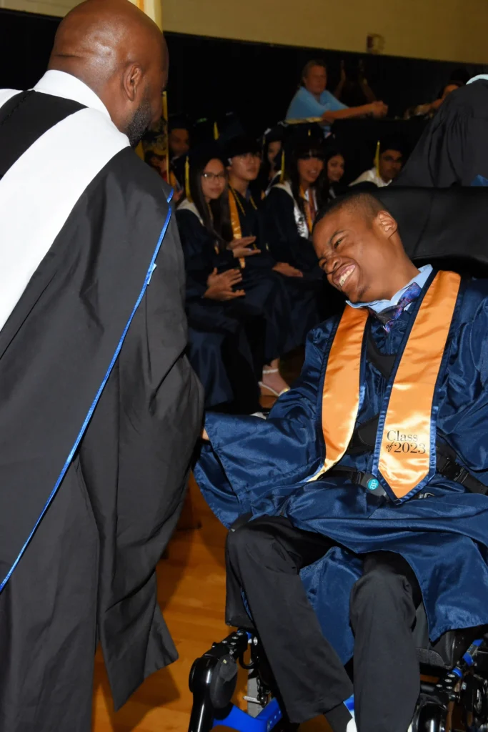 A young man in a wheelchair wearing a navy blue graduation gown with an orange stole labeled "Class of 2023" shakes hands with a faculty member in academic regalia. He has a joyful smile as other graduates in caps and gowns watch in the background.