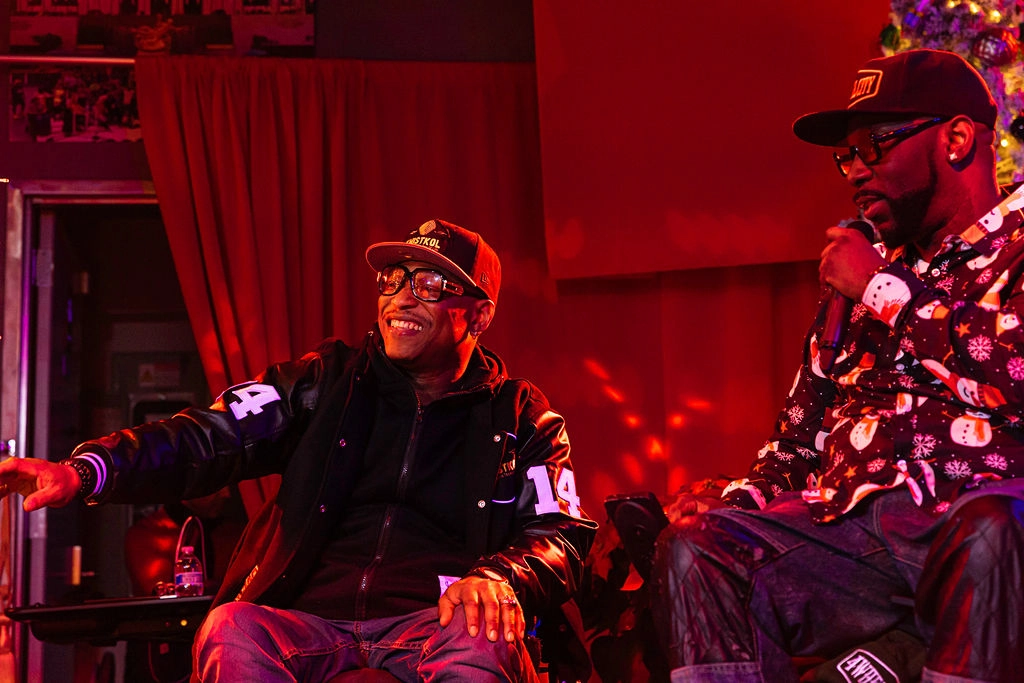 Two men on stage engaged in a conversation. The man on the left is wearing a black baseball cap, glasses, and a black varsity jacket with the number "14" on the sleeve, smiling and gesturing with his right hand. The man on the right is wearing a black cap, glasses, and a festive holiday-themed shirt with snowmen patterns, holding a microphone and speaking. The background is dimly lit with red lighting, a curtain, and a decorated Christmas tree.