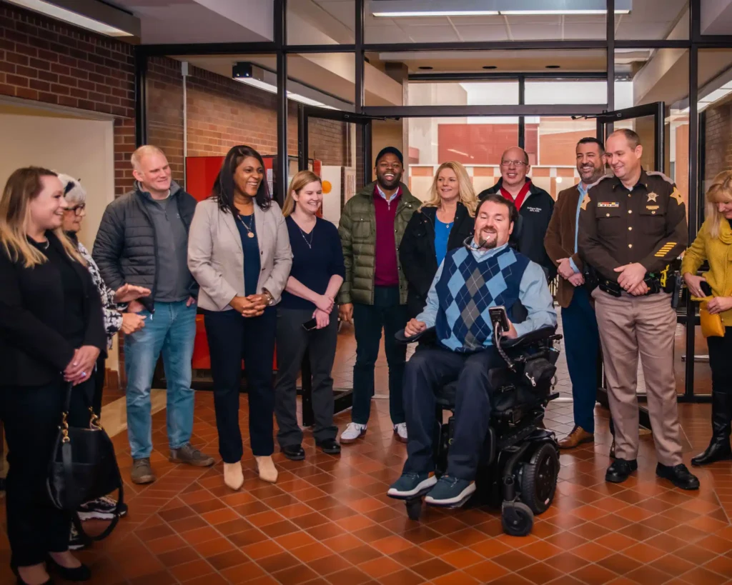 A group of people gathers inside a building, standing in a semi-circle and smiling. A man in a power wheelchair, wearing an argyle sweater, is at the center, holding a device. The group consists of individuals in business and casual attire, including a sheriff in uniform, suggesting a formal or community-related event.