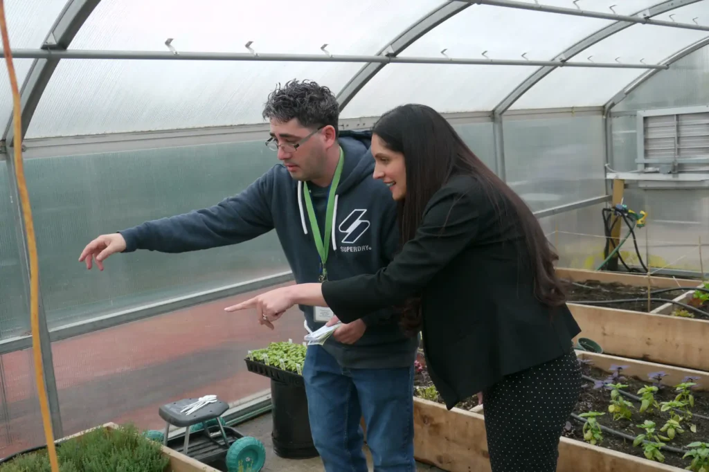 A man wearing a hoodie and glasses is standing inside a greenhouse, pointing towards a section of the raised garden beds, while a woman in a black blazer and polka-dot pants enthusiastically points in the same direction. The greenhouse contains wooden raised beds with young plants growing.