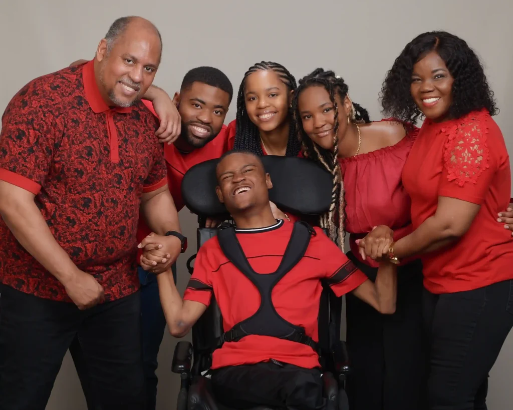 A happy family of six poses together, all wearing red outfits. The young man in the wheelchair is at the center, smiling, as family members hold his hands and lean in affectionately.