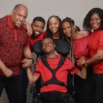A happy family of six poses together, all wearing red outfits. The young man in the wheelchair is at the center, smiling, as family members hold his hands and lean in affectionately.