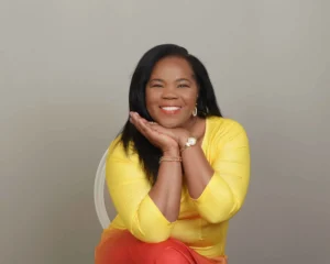 A woman in a bright yellow blouse and orange pants sits gracefully, resting her chin on her hands with a warm smile.