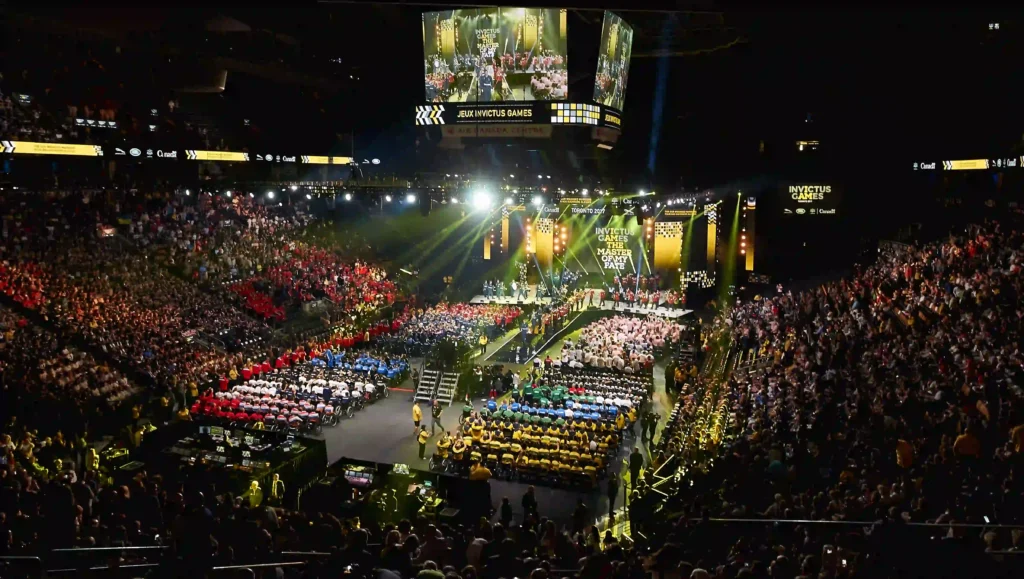 This image shows a large indoor stadium filled with a crowd, with many people seated in color-coordinated sections. The center of the arena has a stage with bright lighting, and large screens displaying the event. The banners and screen text indicate that this is the Invictus Games Toronto 2017, an international adaptive sporting event for wounded, injured, and sick service personnel and veterans. The atmosphere appears grand and celebratory, with participants and audience members engaged in the ceremony. Photo Credit to PRP Productions Toronto 2017