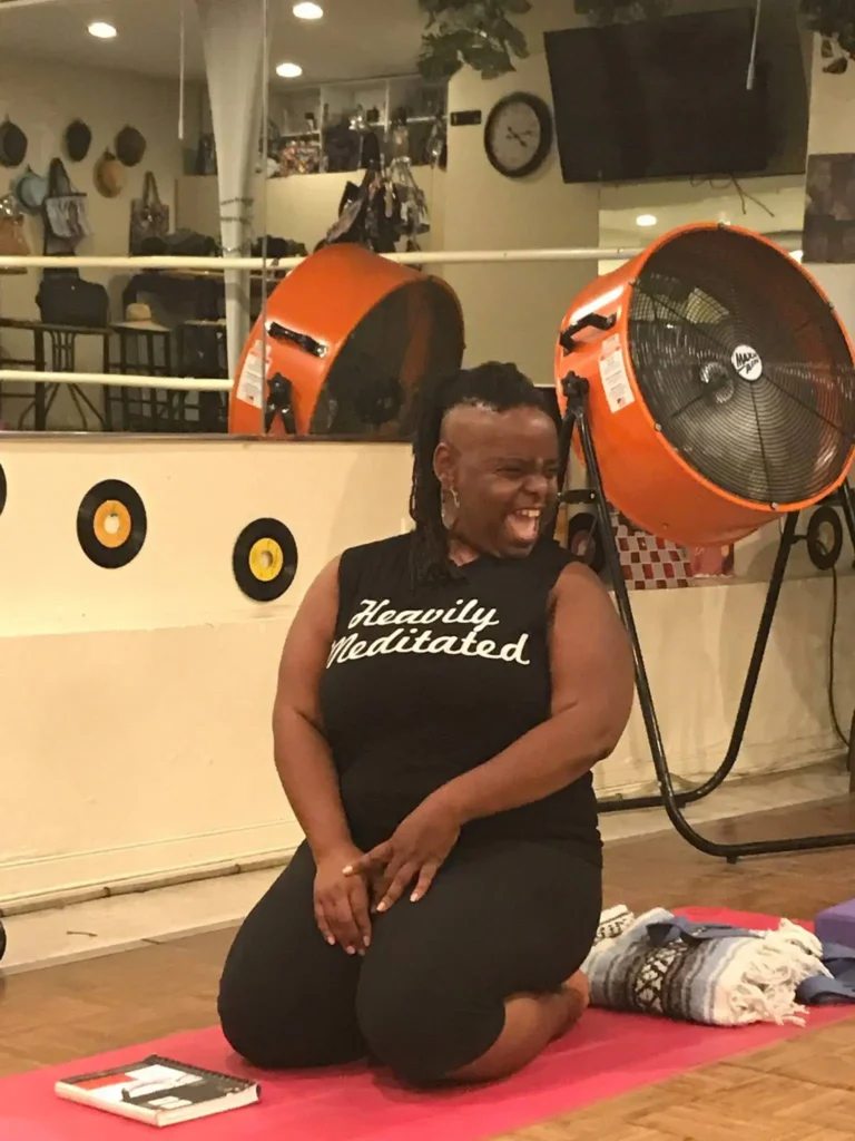 A woman with dark skin and a joyful expression is kneeling on a yoga mat in a room with a mirror and decorative vinyl records on the wall. She is wearing a black sleeveless shirt with the phrase "Heavily Meditated" written in white cursive. Her hair is styled with shaved sides and long locs. Large orange industrial fans are visible in the background, along with yoga props such as a blanket and blocks.