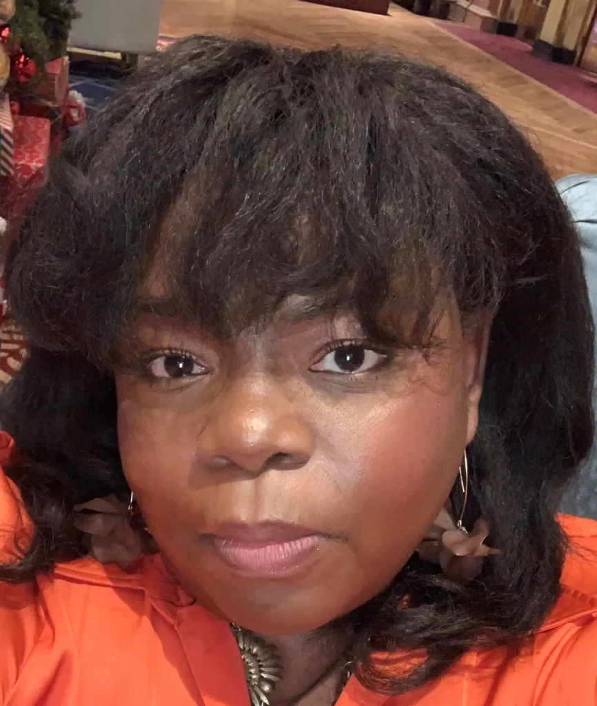 A close-up selfie of a woman with dark skin, dark eyes, and full lips. She has a neutral yet warm expression and is wearing a vibrant orange top with large floral earrings. Her dark, wavy hair frames her face, and the background is slightly blurred with a festive setting, including wrapped gifts and a red carpet.
