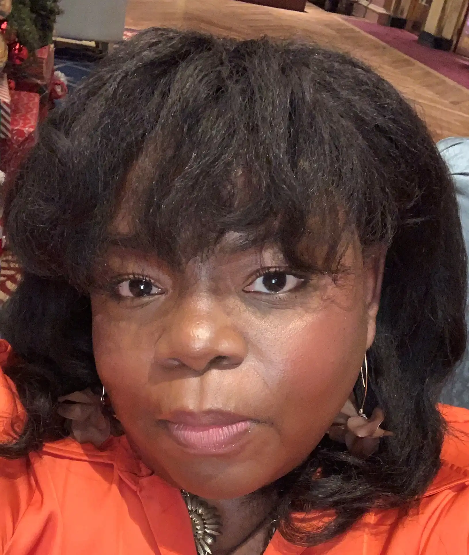 A close-up selfie of a woman with dark skin, dark eyes, and full lips. She has a neutral yet warm expression and is wearing a vibrant orange top with large floral earrings. Her dark, wavy hair frames her face, and the background is slightly blurred with a festive setting, including wrapped gifts and a red carpet.