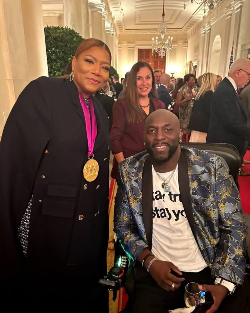 Queen Latifah, wearing a gold medal on a pink ribbon and a stylish black outfit, poses with a smiling man in a wheelchair who is dressed in a blue and gold floral blazer over a white t-shirt with black text. They are at a formal event in a grand hallway with chandeliers, high ceilings, and a crowd of elegantly dressed attendees engaged in conversation