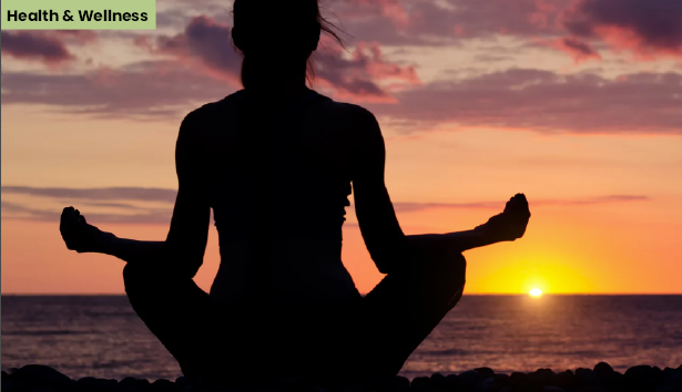 Silhouette of a person practicing yoga in a meditative pose by the ocean at sunset. The sun sets on the horizon, casting a warm glow over the sky and water. A 'Health & Wellness' label is positioned in the top left corner.