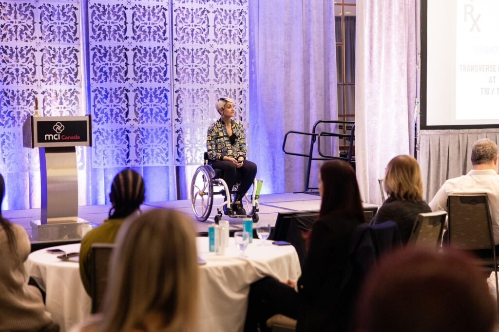 A woman in a wheelchair is giving a presentation on stage at a professional event. She is holding a microphone and appears to be engaging with the audience. The stage has a decorative patterned backdrop with blue lighting. The podium bears a sign reading "MCI Canada," and an audience is seated at round tables, attentively listening.
