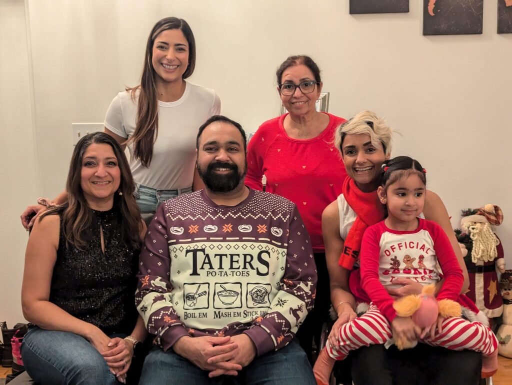 A group of six people, including a young child, is posing for a festive holiday photo indoors. They are all smiling and dressed in casual and holiday-themed attire. The man in the center is wearing a sweater with the words "TATERS PO-TA-TOES" and illustrations referencing "boil 'em, mash 'em, stick 'em in a stew." The group includes individuals of different ages, and some are wearing red and white festive clothing. The background includes Christmas decorations.