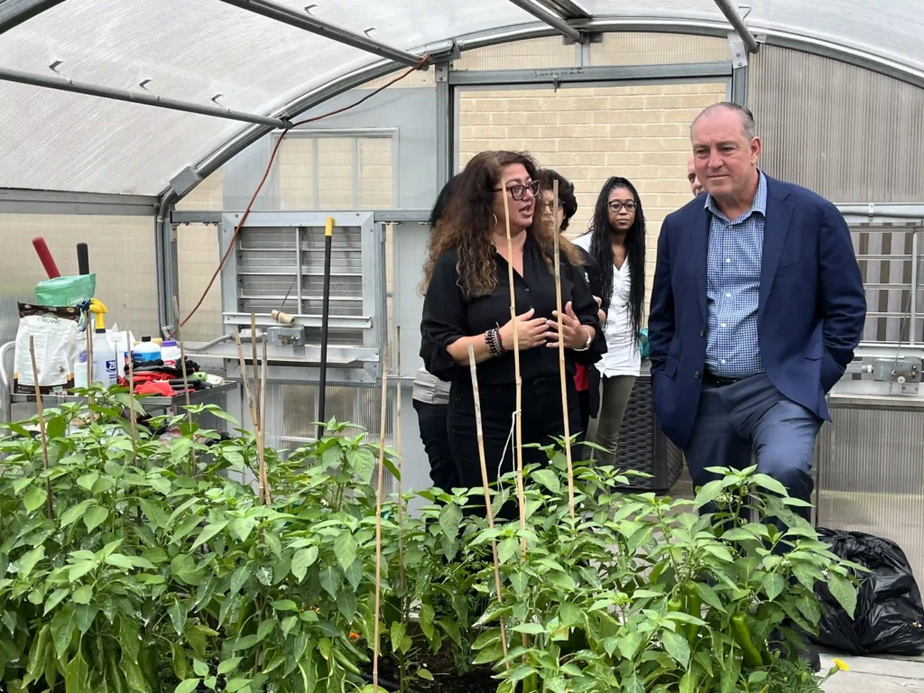 A woman with curly hair, wearing a black blouse, is speaking and gesturing with her hands while standing inside a greenhouse. A man in a navy blue suit with a blue shirt listens with a slight smile, leaning slightly against the raised garden bed. Several people stand in the background, and there are green plants growing in raised beds with wooden supports.