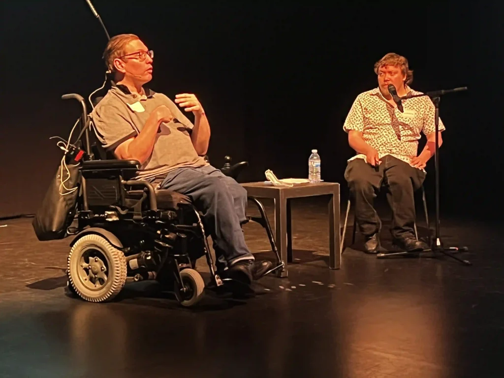 Two individuals on a dimly lit stage engaged in a discussion. One person, wearing glasses and a gray shirt, is seated in a power wheelchair and using sign language. The other person, in a white patterned shirt, is seated on a chair, speaking into a microphone. A small table with water bottles and papers is placed between them.