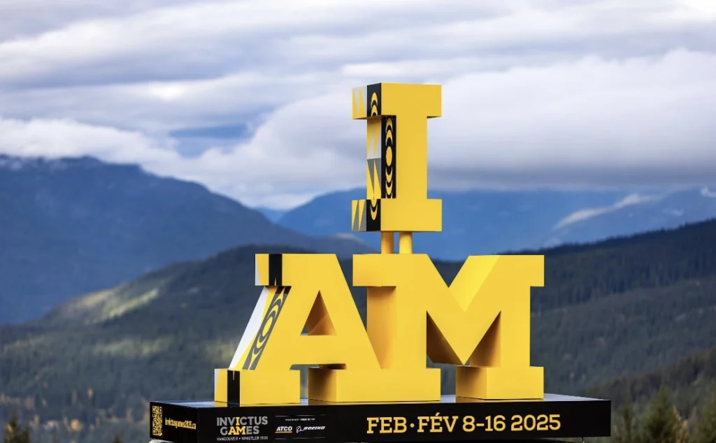 A large yellow 3D sculpture spelling 'I AM' is displayed against a mountainous backdrop with cloudy skies. The structure features Indigenous-inspired black and white patterns on parts of the letters. The base of the sculpture includes text indicating the 'Invictus Games Vancouver-Whistler 2025,' scheduled for February 8-16, along with sponsor logos for ATCO and Boeing. Photo Credit: Jeremy Allen