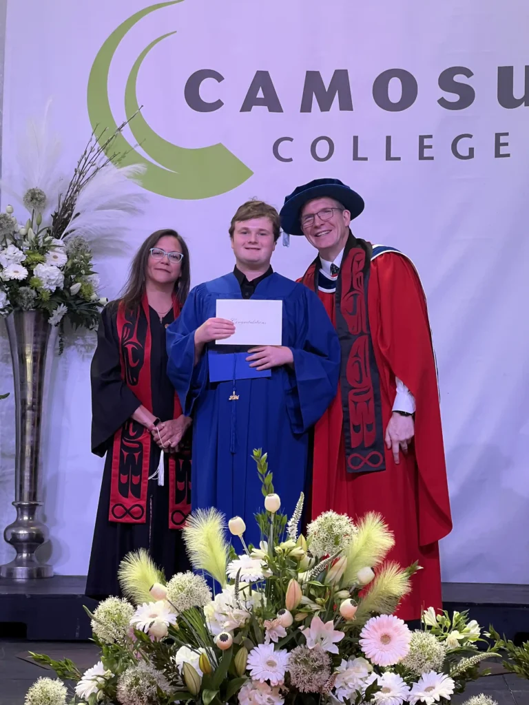 A young graduate in a blue gown holds a certificate while standing between two academic staff members dressed in traditional regalia. They are on a stage with a large "Camosun College" banner in the background, surrounded by floral decorations. The young graduate has a proud expression as they celebrate their achievement.