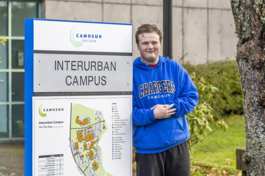 A young man wearing a blue "Camosun Chargers" hoodie stands beside a sign that reads "Camosun College Interurban Campus." He is smiling and posing casually with his arms crossed in front of a tree and a campus map. The background includes a building and green landscaping.