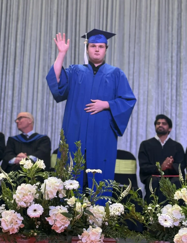 A young graduate in a blue cap and gown stands on a stage with a backdrop of gray curtains. He raises his right hand in a formal gesture while placing his left hand on his chest. There are floral decorations in front of him, and faculty members in academic regalia are seated behind him, some clapping.