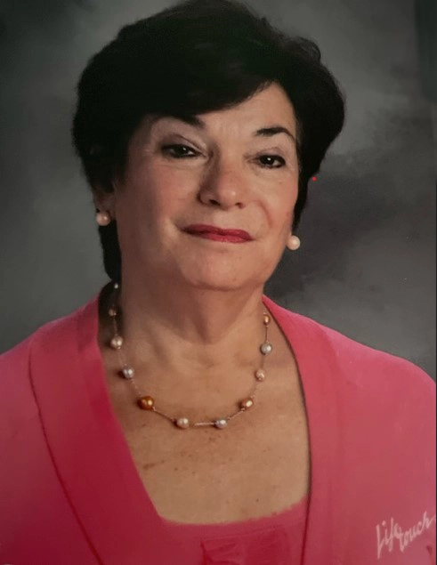 A professional portrait of Jane Fischer, an older woman with short dark hair, wearing a pink blazer over a matching top and a pearl necklace. She has a warm yet serious expression, and the background is a simple, neutral tone.