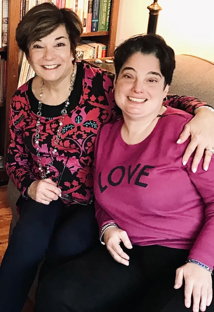 Two smiling women sitting together in a cozy indoor setting with bookshelves in the background. The woman on the left wears a red and black patterned top with a long necklace, and the woman on the right wears a pink sweater with the word "LOVE" written on it. They are sitting close, with one arm wrapped around each other.