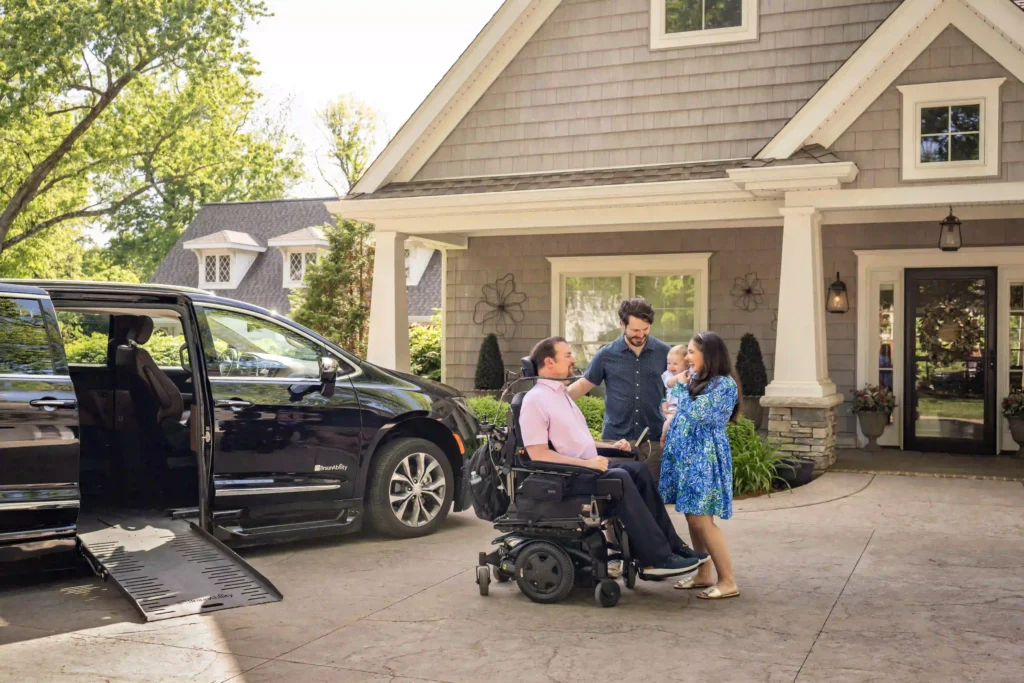 A man in a power wheelchair is outside a suburban home, conversing with a smiling couple and their baby. A black wheelchair-accessible van with an extended ramp is parked in the driveway. The setting is bright and inviting, with lush greenery surrounding the home.