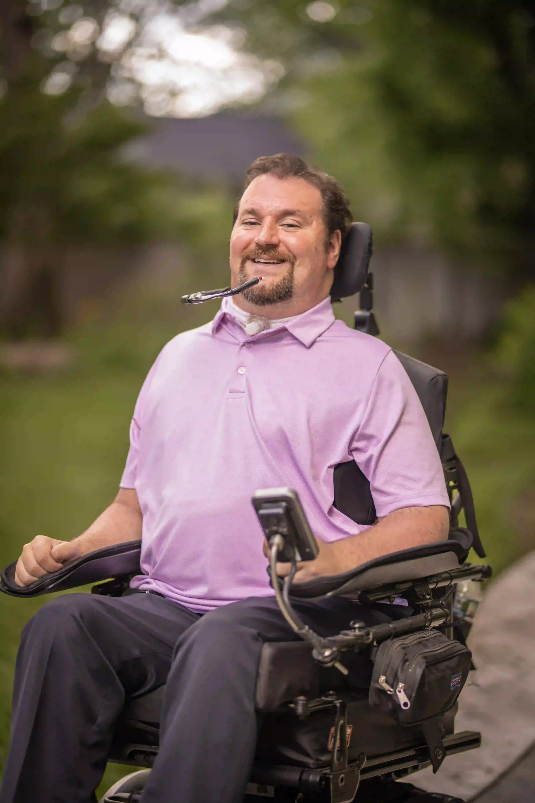 A man with a beard, wearing a light purple polo shirt, sits in a power wheelchair outdoors. He has a joyful expression and uses a mouth-controlled device to operate his wheelchair. The background is blurred with greenery